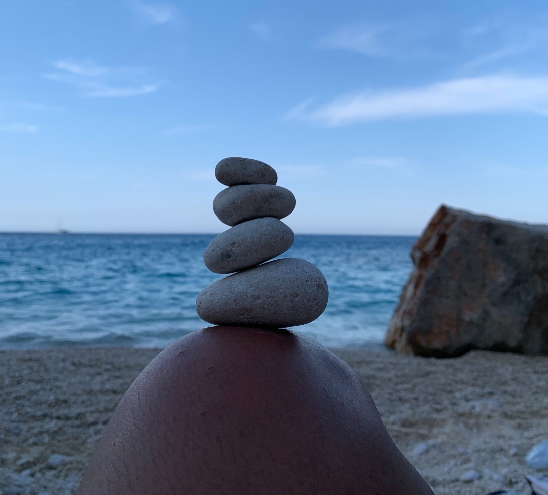 pedras de diferentes tamanhos em equilíbrio sobre um joelho. Vê-se o mar azul ao fundo.
